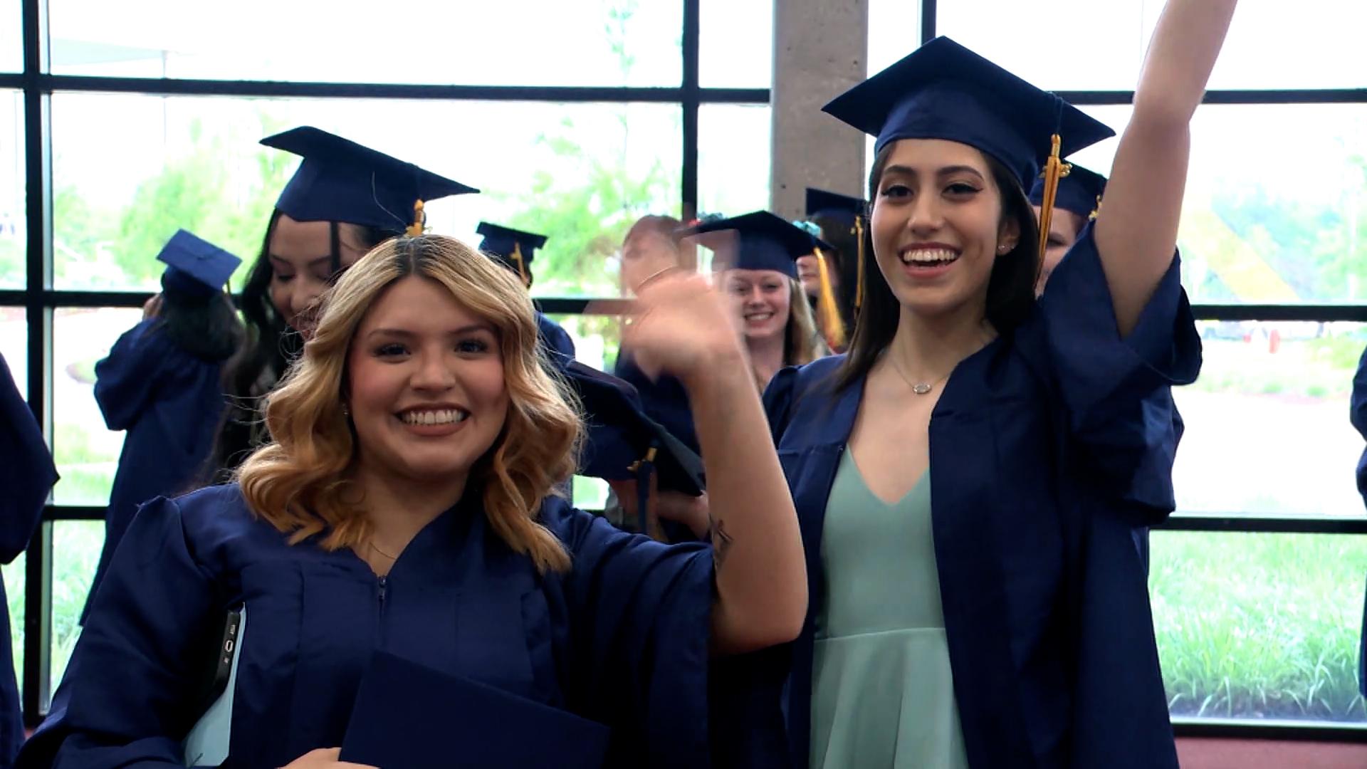 Two graduates in caps and gowns waving at the camera.